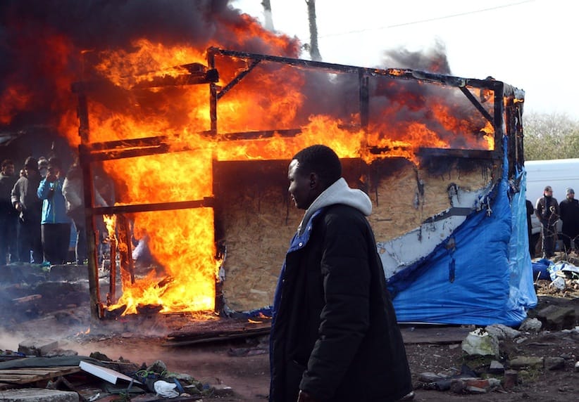 A migrant walks past a burning dwelling in the makeshift migrants camp near Calais, France in March, 2016. A destruction crew on Wednesday, March 16 tore down the last of more than 1,000 tents and huts in a large swath of a makeshift migrant camp in the French port city of Calais, a job marked by fiery protests, an ongoing hunger strike and several arrests. The prefecture, or state authority, which ordered the dismantling of the southern sector of the camp - a veritable village on 7.5 hectares (18.5 acres) of land - said workers guarded by police finished pulling down the flimsy dwellings at 2 p.m. Wednesday, less than three weeks after the job began. (AP Photo/Michel Spingler, file)