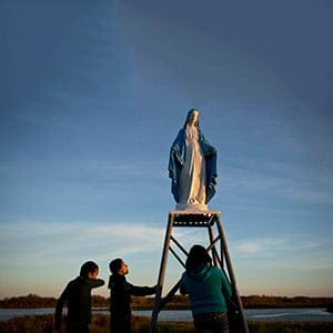 Attawapiskat, Ontario, Canada. Young people play at a statue of the Virgin Mary near the St. Francois-xavier Catholic Church on the shores of the Attawapiskat river. The community has a complicated history with the Catholic church with many of its members subjected to horrible abuses while at the Catholic run St. Anne's Residential school. A conversation with one 59 year old man who had endured St Annes for six years remained with me. He told me of repeated sexual and physical abuse at the hands of priests and nuns and in one particular instance when he was 11 years a nun punched him and broke his nose. It was the last time in his life that he was able to cry. To this day he still carries deep shame and wishes he could express it through tears, however even at the funerals of his parents and his own daughter, he was unable to. At the same time many in the community still connect to their Catholic faith, a testament to their forgiveness. October 7, 2016. David Maurice Smith/Oculi for The Pulitzer Center on Crisis Reporting.