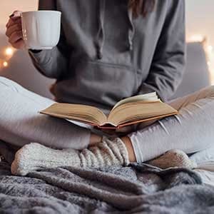 Girl holding cup of hot tea and reading in bed. Around her in bad earphones, book, smart phone. Decorative lights in background.