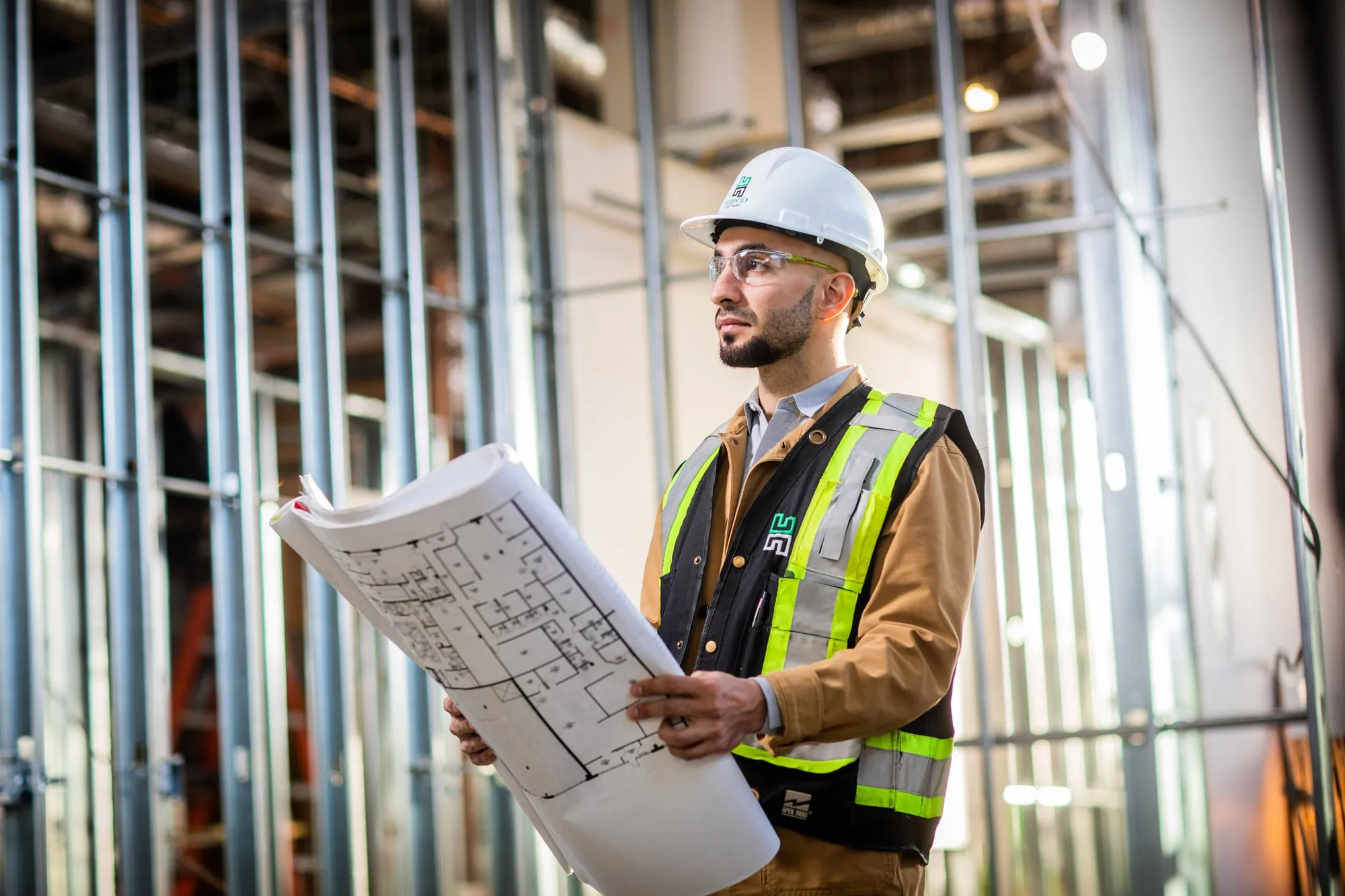 A man in a white construction hat and hi-vis vest holding construction drawings.
