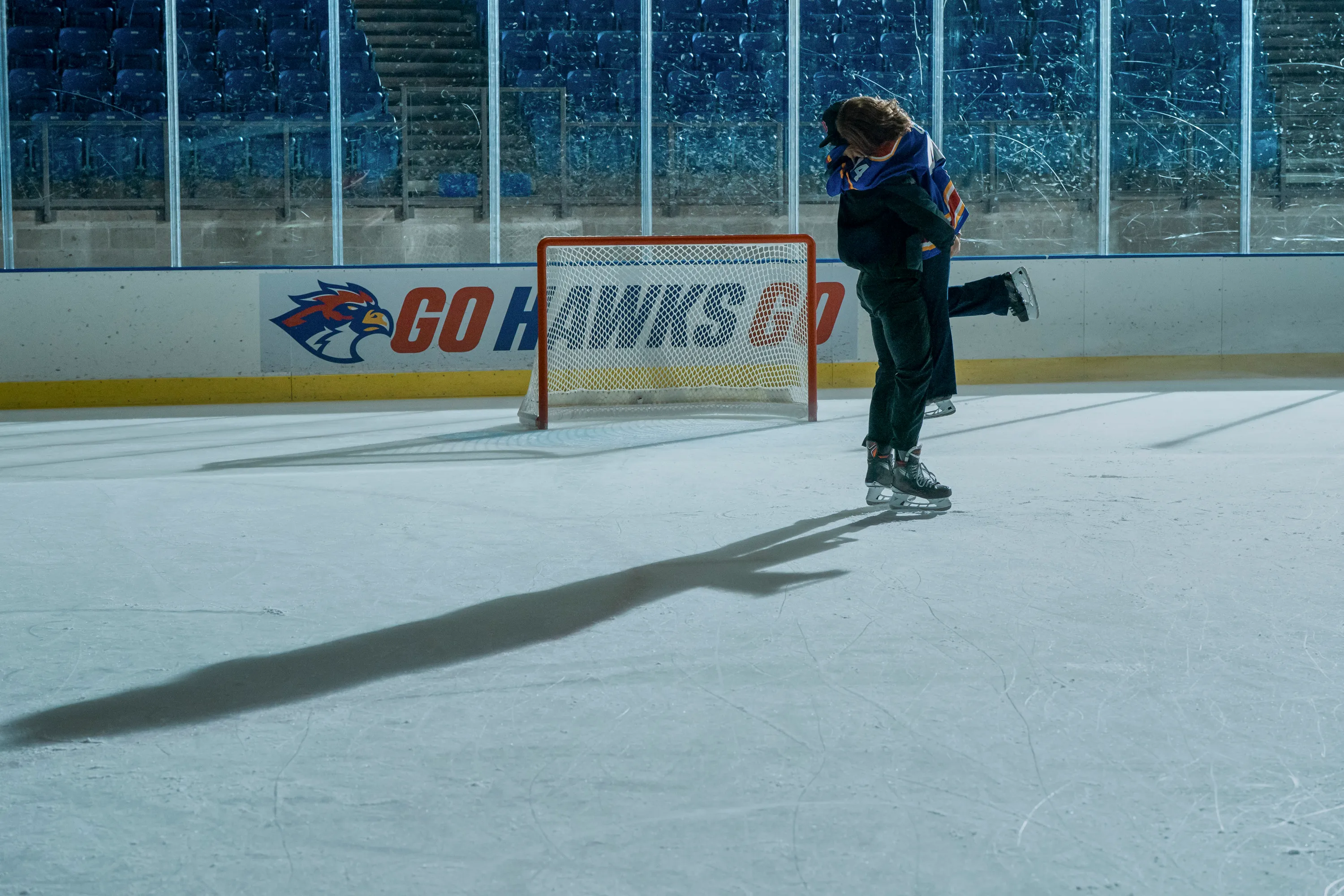 Two people in hockey uniforms and skate shoes are hugging each other on a hockey link.