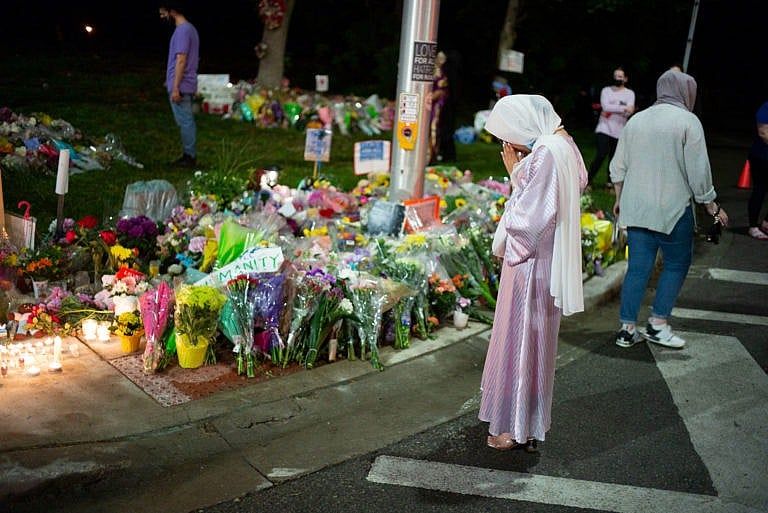 A woman in a purple dress and white headscarf stands in front of a makeshift memorial with many flower bouquets placed on the ground