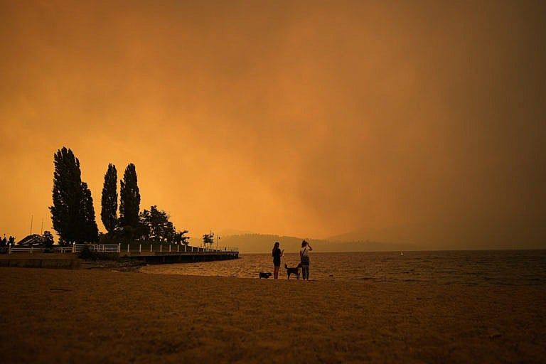 Two people stand on a beach looking at glowing orange smoke across a lake.