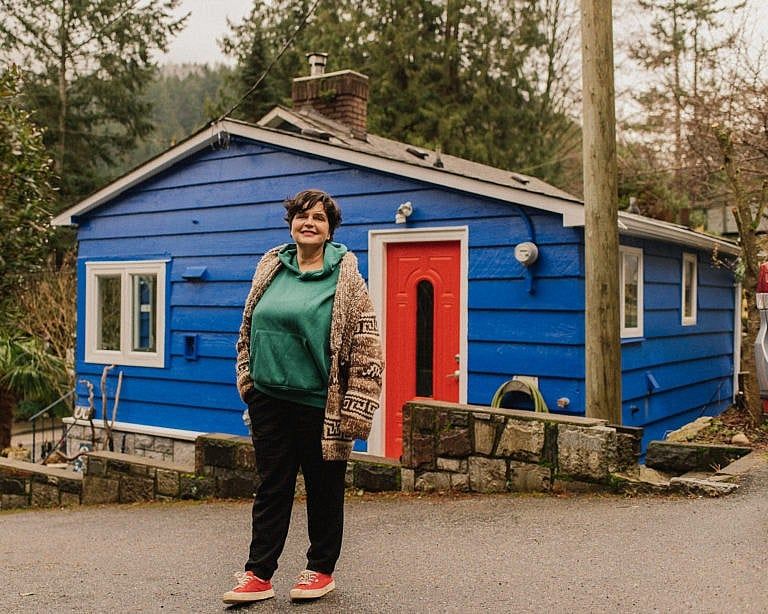A photo of a woman with a pixie cut, wearing a green hoodie, brown cardigan, black pants, and red sneakers. She's standing in front of a small blue cottage with a red door.