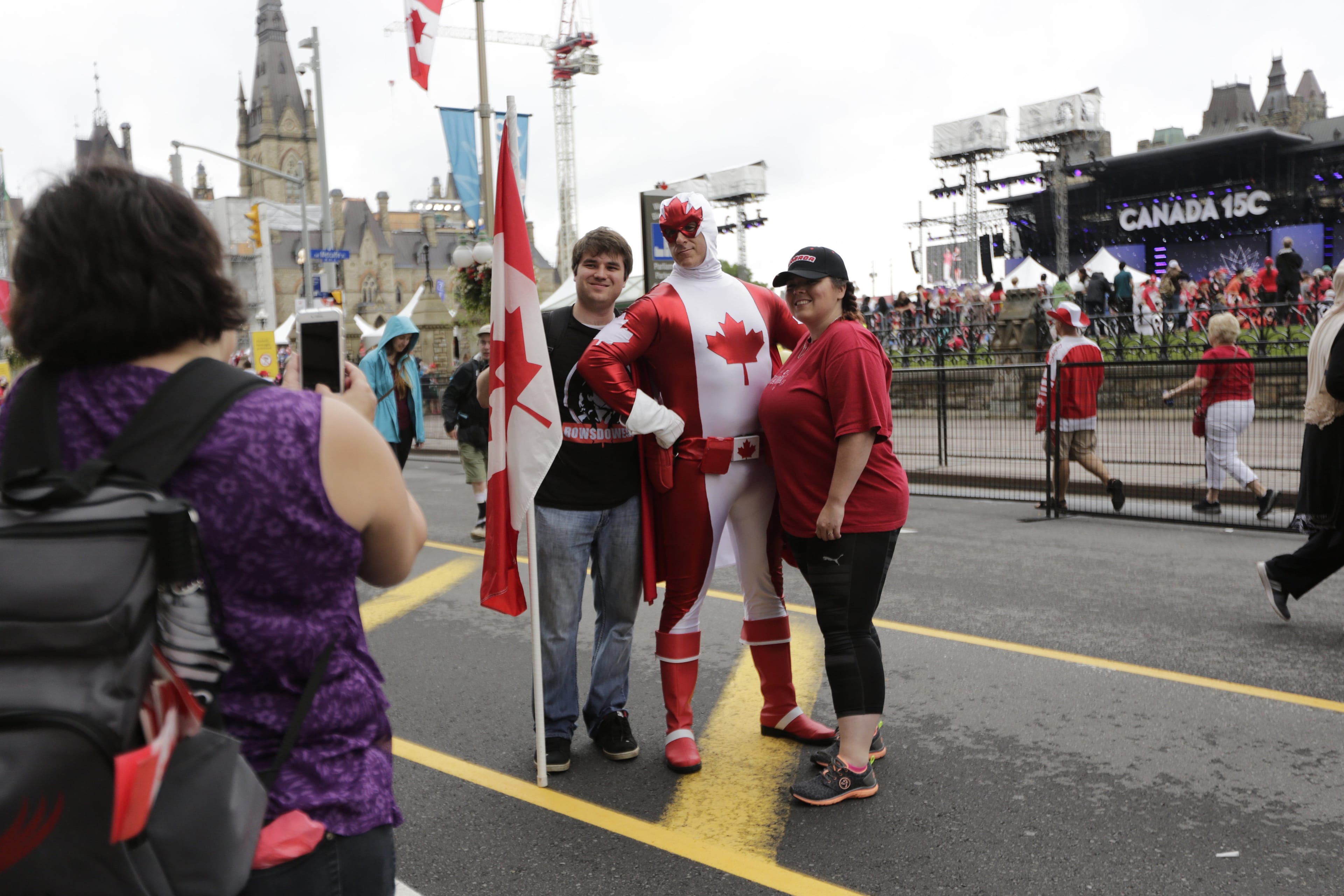 Attendees stand for a photograph with a performer dressed as "Captain Canada" during the Canada Day event on Parliament Hill in Ottawa, Ontario, Canada, on Saturday, July 1, 2017. The 150th anniversary of Canada, promoted as Canada 150, marks the sesquicentennial anniversary of the Canadian Confederation. Photographer: David Kawai/Bloomberg via Getty Images