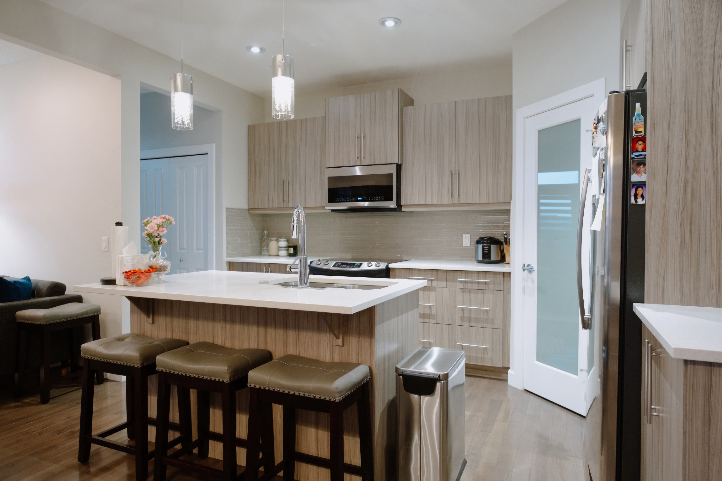 A brightly lit kitchen with an island in the middle, 3 chairs against its side and a white door in the corner.