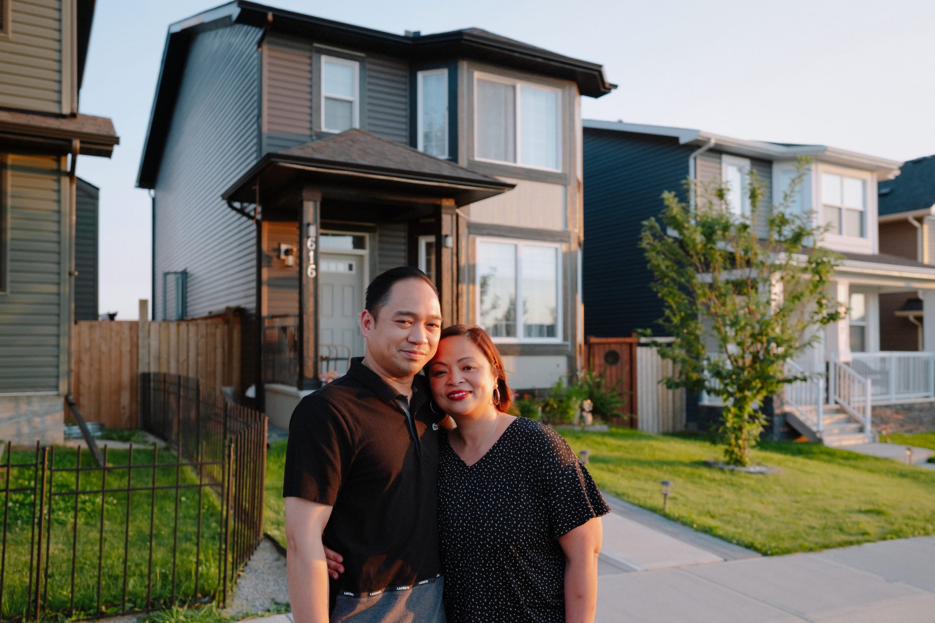 A man and woman stand in front of their house, giving each other a side hug