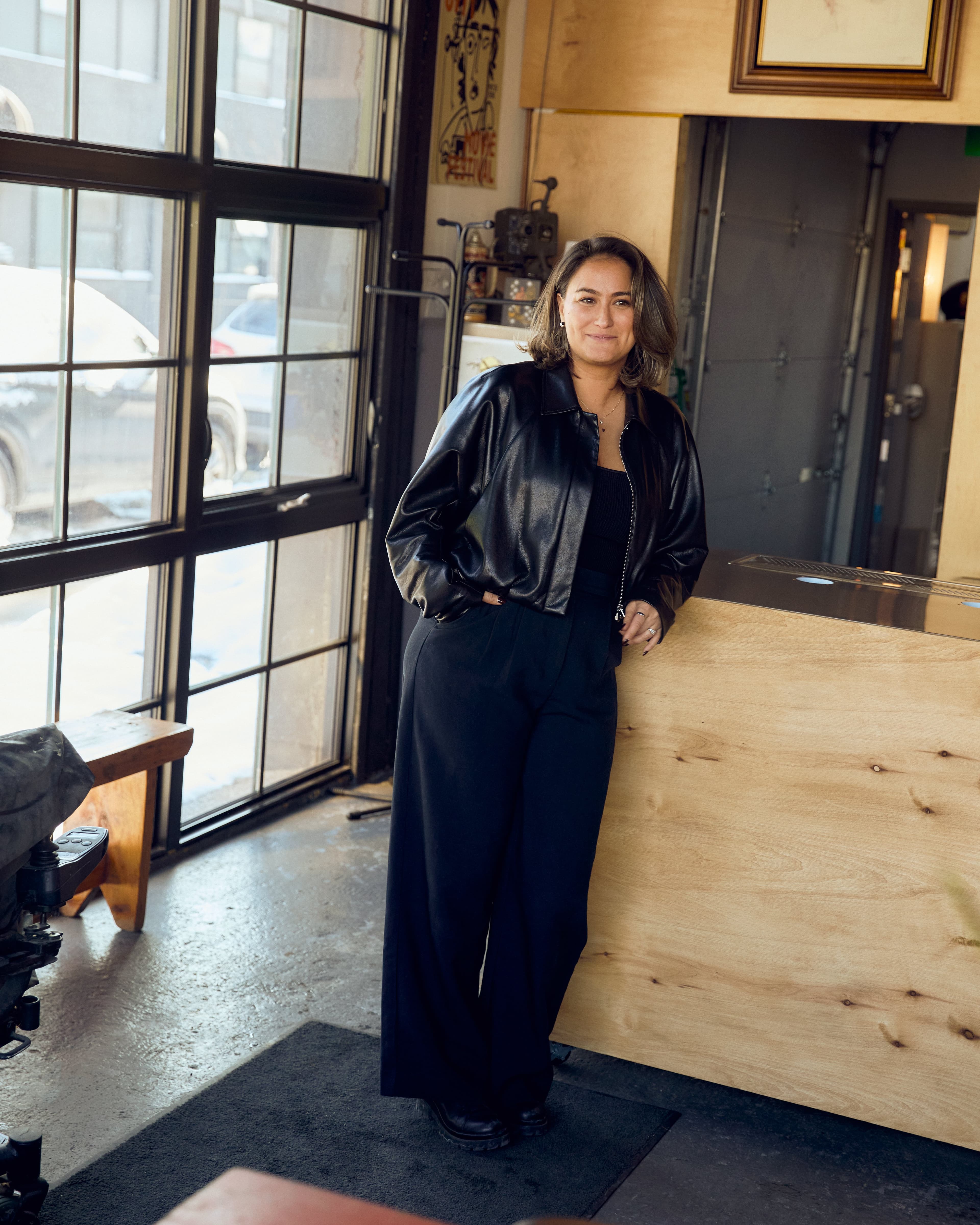 A woman in a leather jacket and wide-leg pants standing in front of a reception desk in an office.