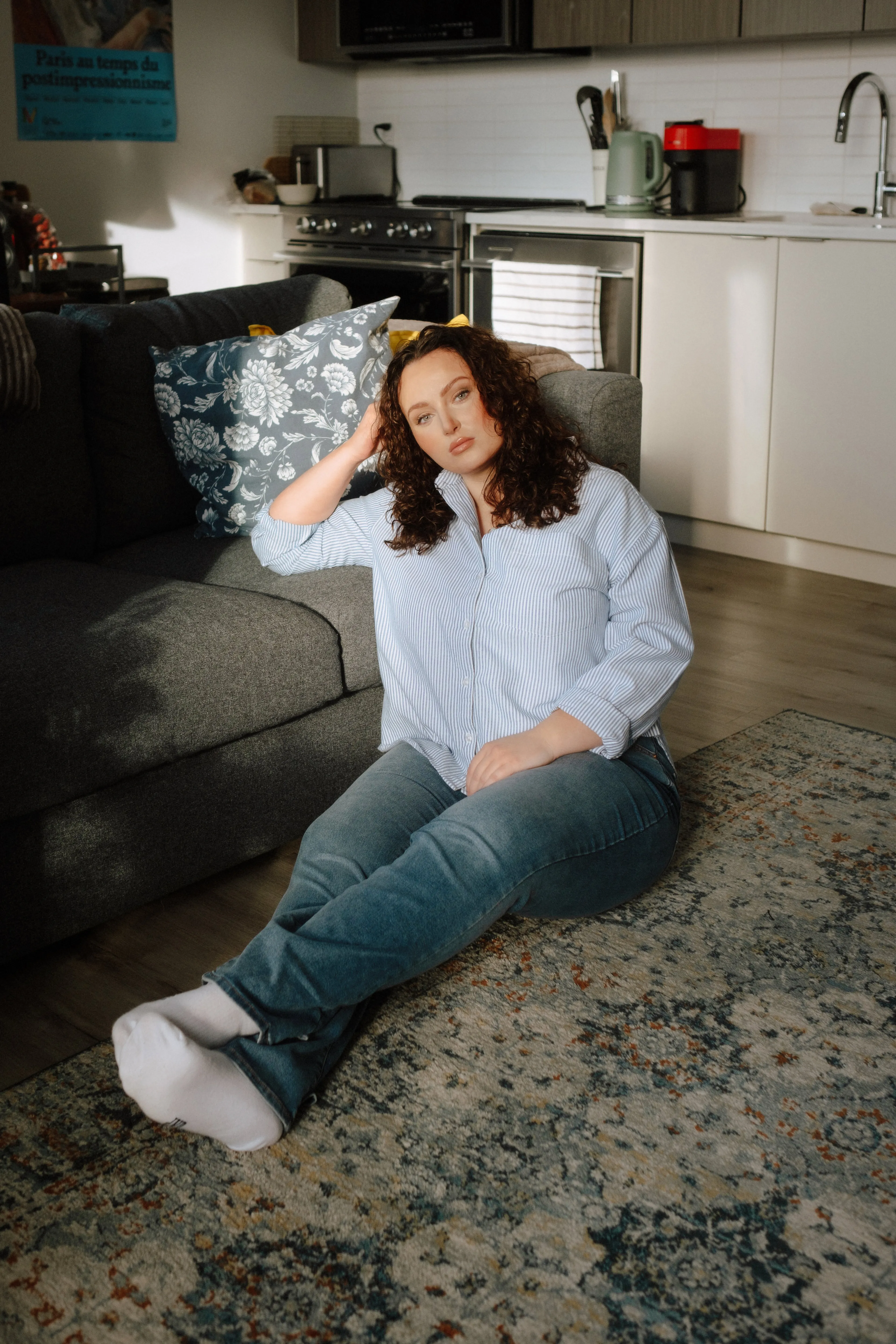A woman sitting on the floor laying against a couch atop a rug in an apartment
