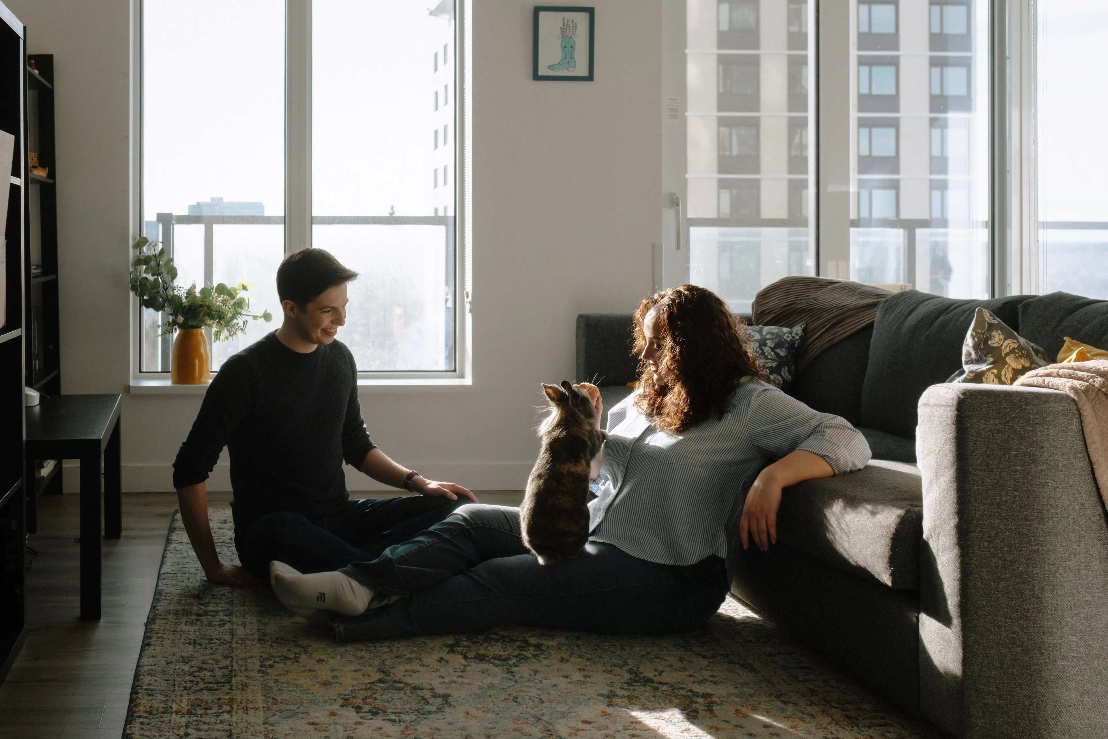 A woman and man sitting on the floor of their living room in a high-rise apartment. Other apartment buildings can be seen outside two windows