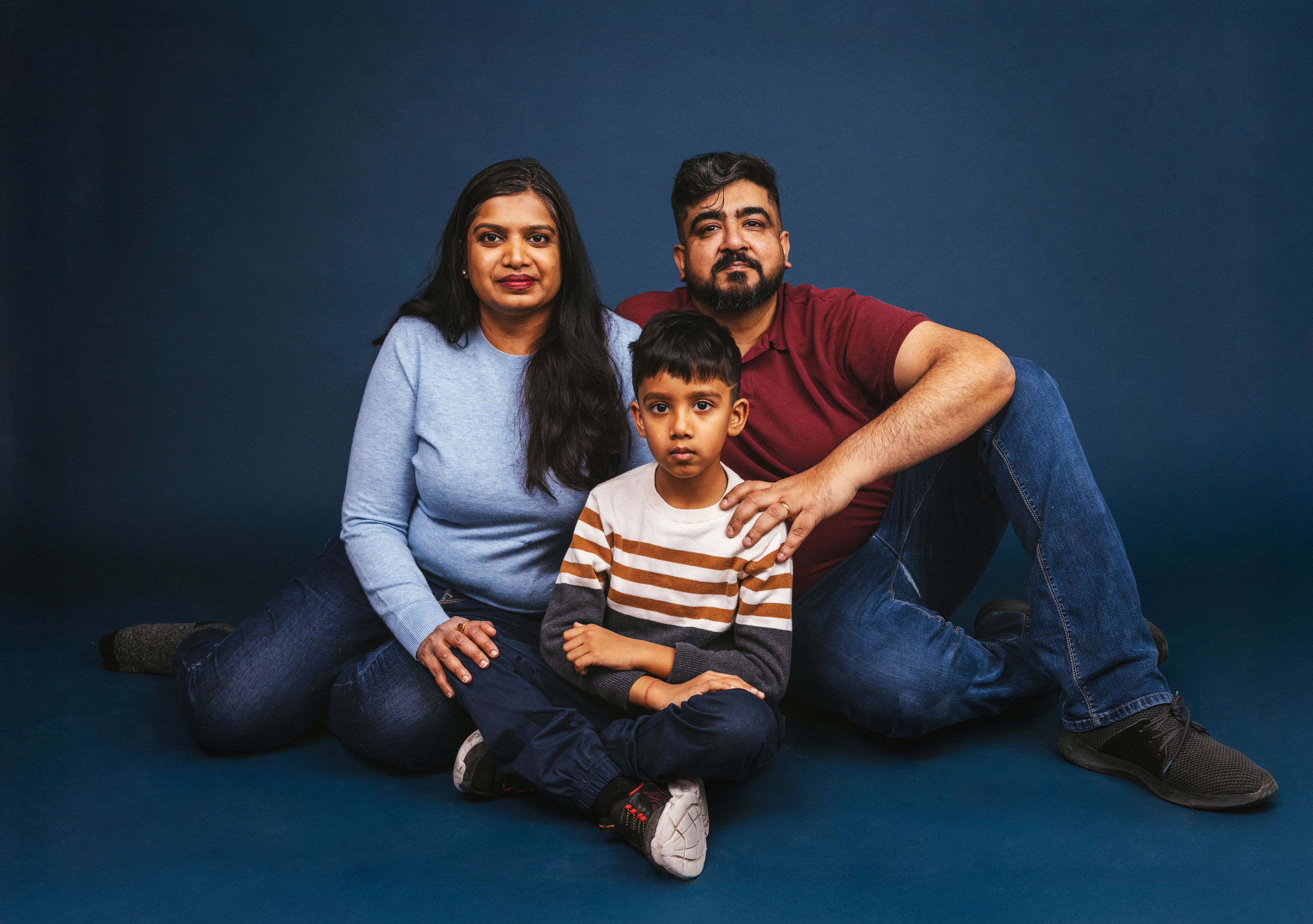 A portrait of a family sitting on the ground. A man wearing a red golf shirt, with a woman sitting to hid left in a light blue knit sweater and a boy sitting in front go them with his legs crossed, arms in lap.