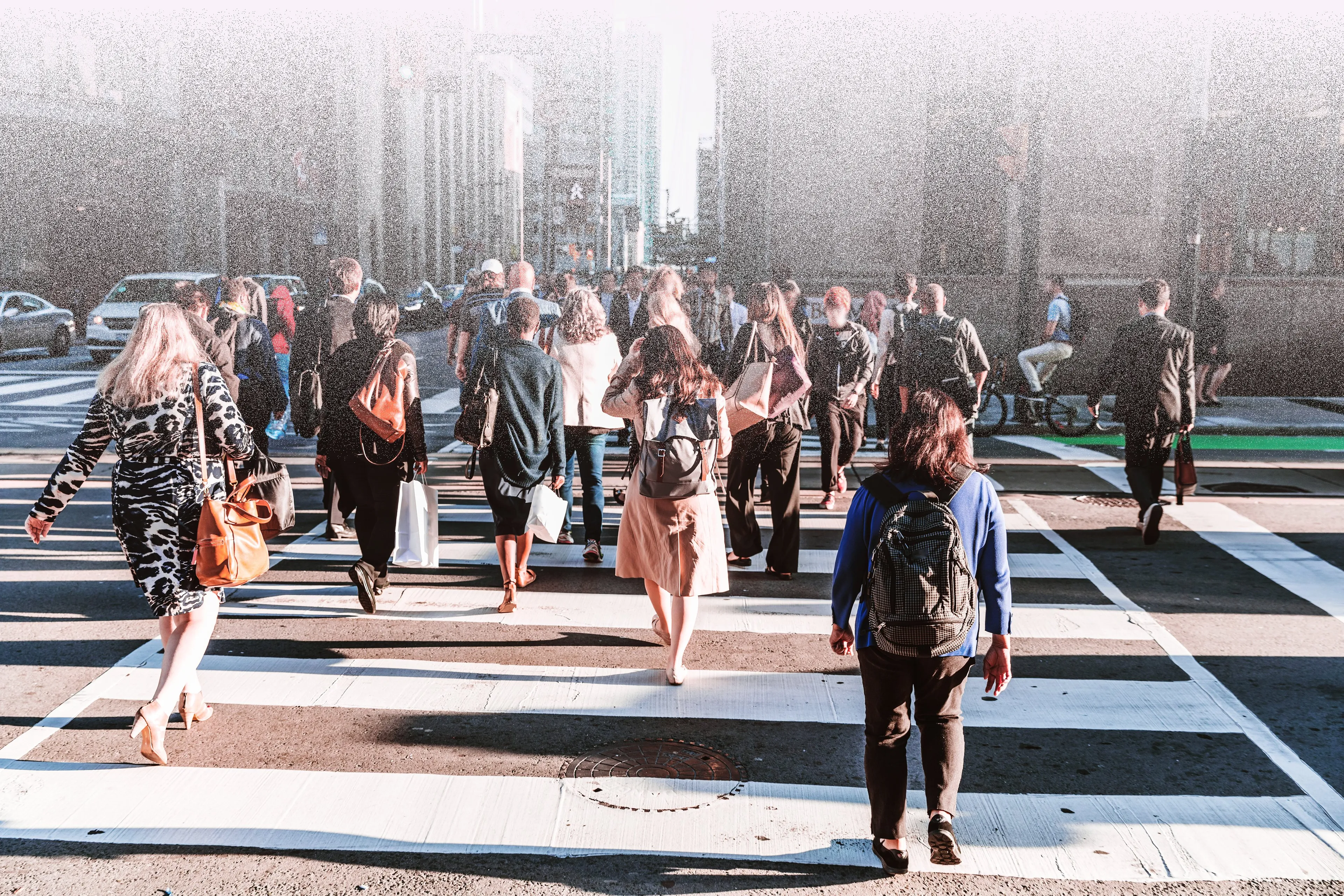 Crowd of pedestrians crossing a sunlit city intersection during rush hour, with long shadows and tall buildings in the background.