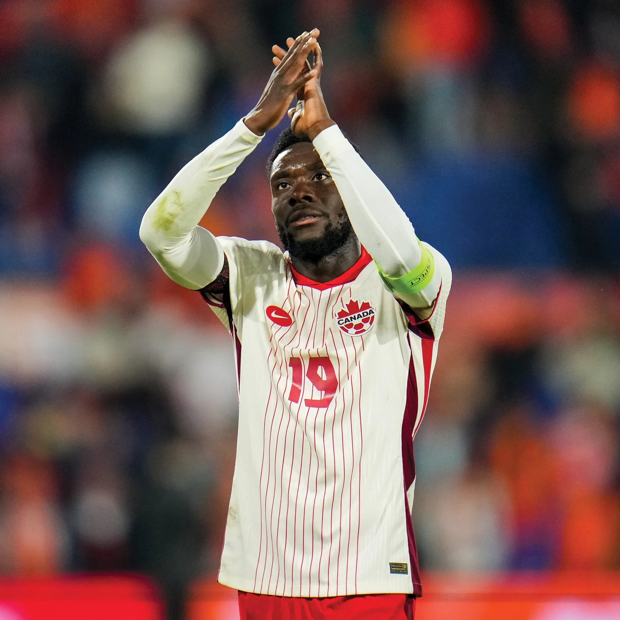 A soccer player (Alphonso Davies) is clapping hands above his head. Behind him is the blurred figures of the audience at the seats