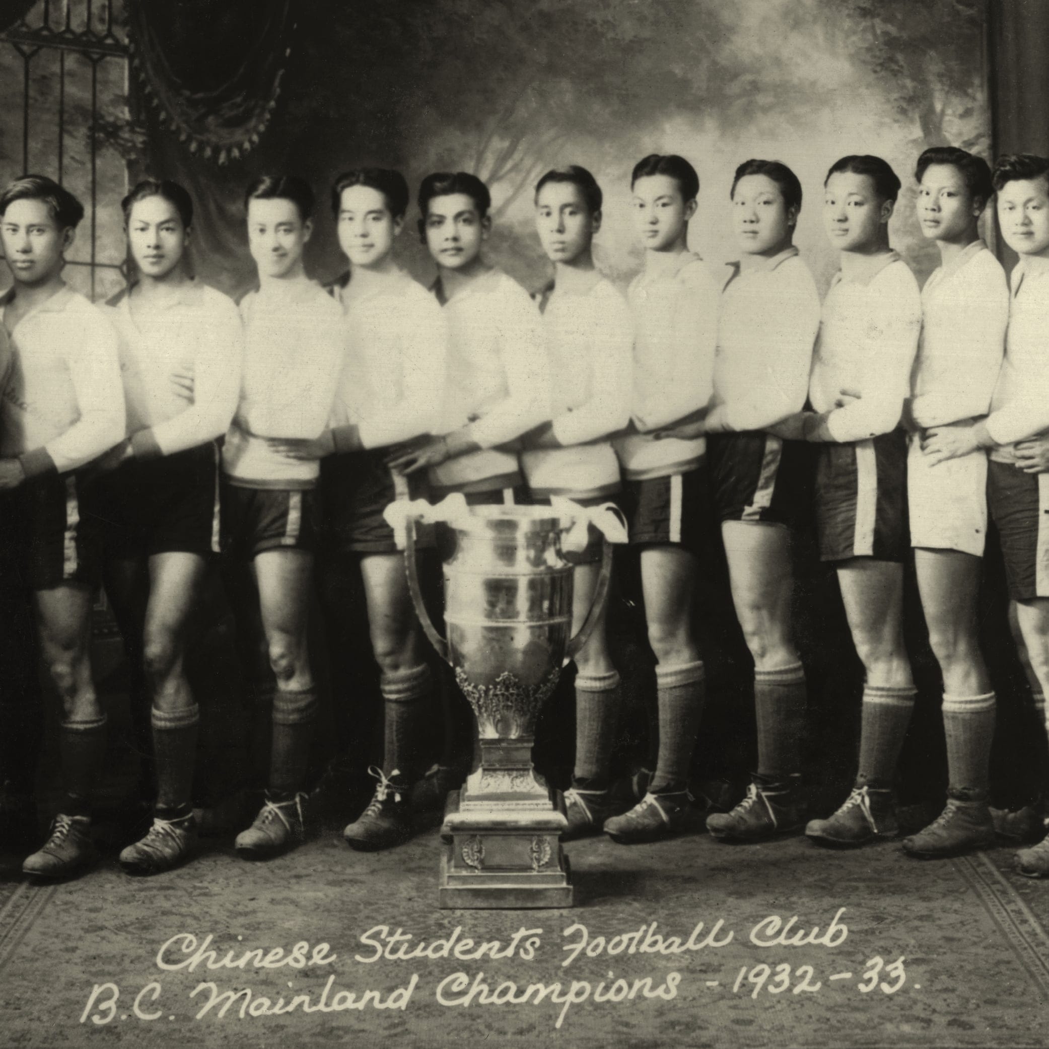 Sepia toned, black-and-white picture in which 15 men are lining up, posing for the picture. A huge, shiny trophy is put in front of them, and under that, a hand-written note says "Chinese students football club B.C. mainland champions - 1982-33."