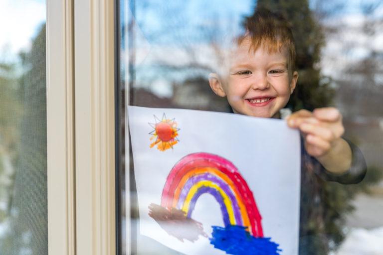 Young Boy taping rainbow drawing on window during the Coronavirus pandemic.