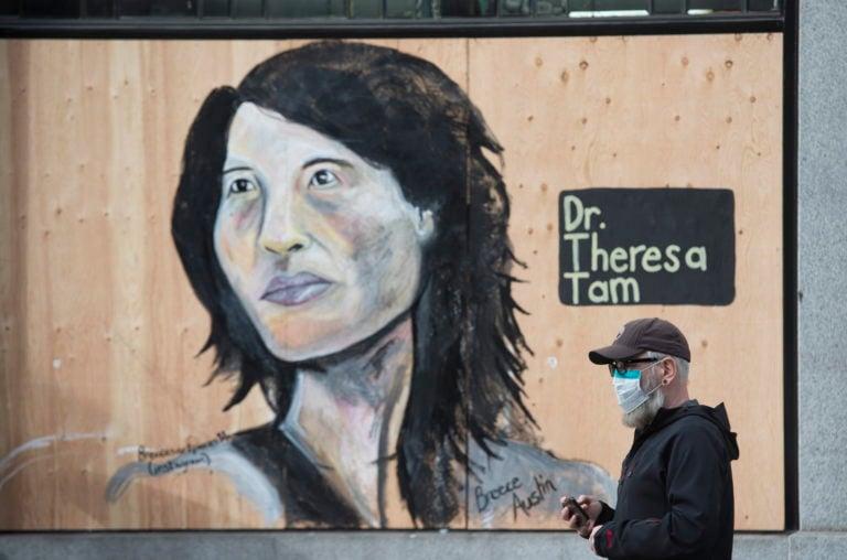 A man wears a protective face mask as he walks past a portrait of Dr. Theresa Tam on a boarded up shop in downtown Vancouver, B.C. on Apr. 1, 2020. (Jonathan Hayward/CP)