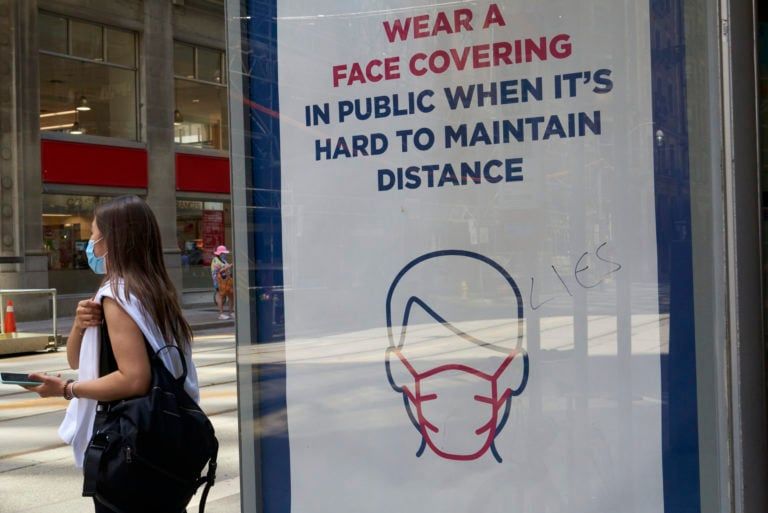 A woman holding a cell phone stands near a vandalized sign reminding Torontonians to wear face coverings in public when it’s hard to maintain distance on Aug. 5, 2020. (Rachel Verbin/CP)