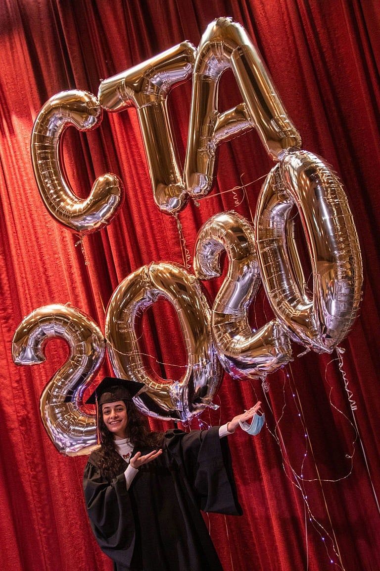 Sham Al Mukdad stands in front of balloons spelling out CTA 2020, wearing a graduation cap and gown