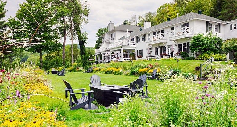 A wide angle shot of a large white house surrounded by greenery and a couple of black patio chairs on the lawn.