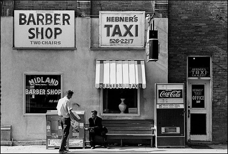 A photo of a storefront with signs reading "BARBER SHOP: TWO CHAIRS" and "HEBNER'S TAXI 526-2217". In front of the storefront are two men in conversation.